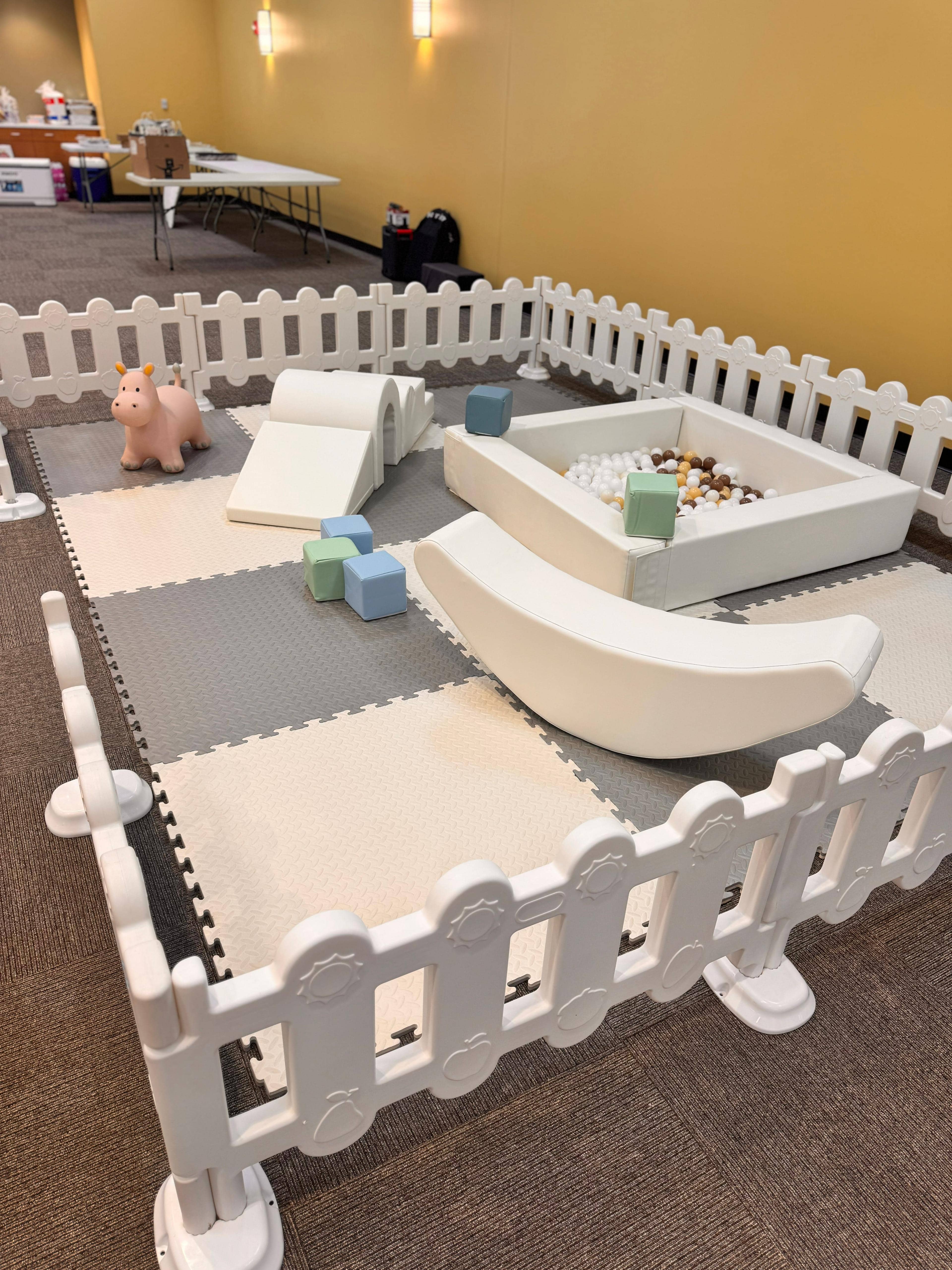 Soft play rental setup indoors — neutral-tone foam mats, ball pit, soft climbers and white safety fence for toddler birthday parties