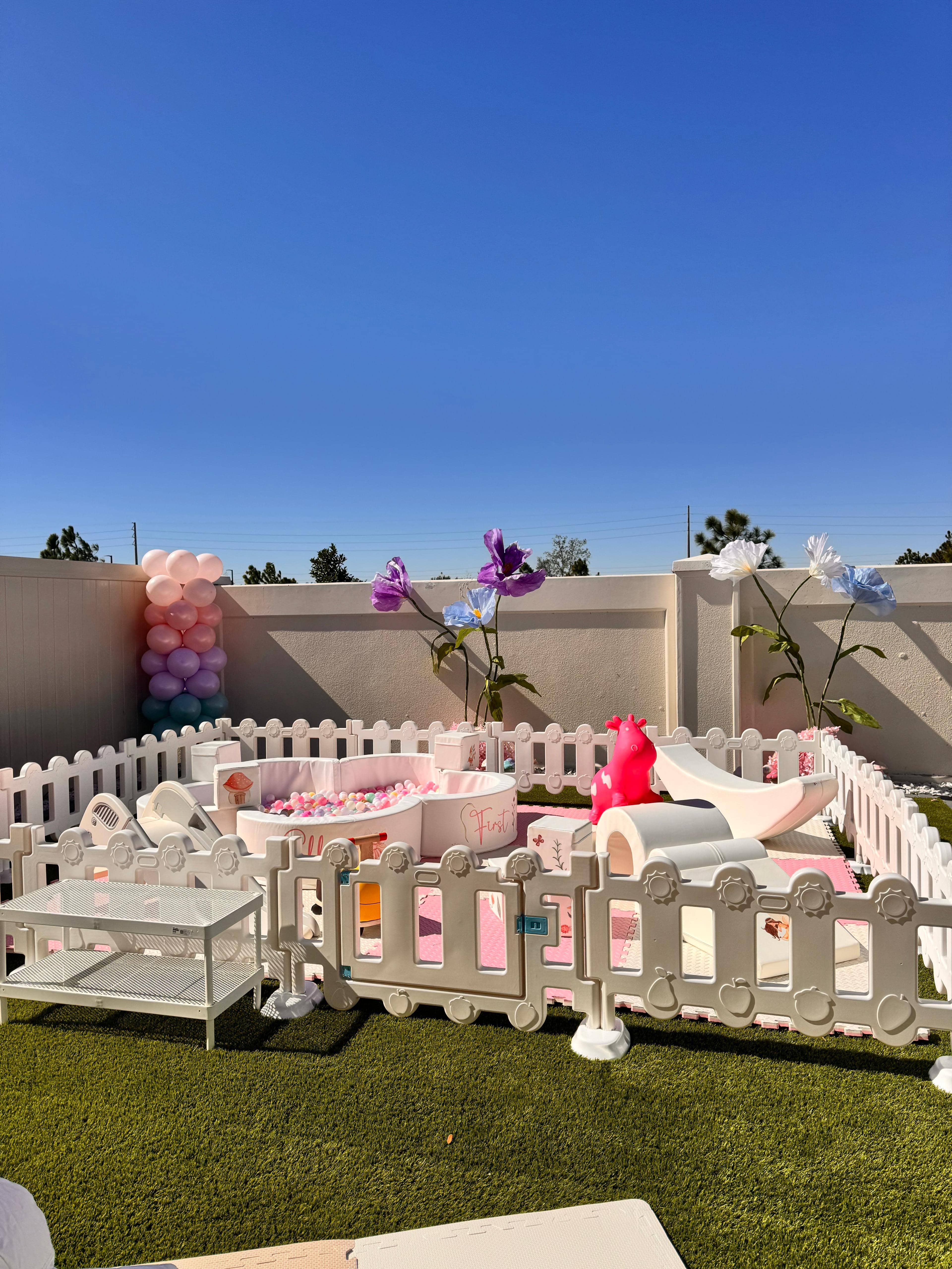 Wide view of Blush and Bloom soft play rental in a backyard — pink foam mats, floral ball pit, slide and balloon column decoration